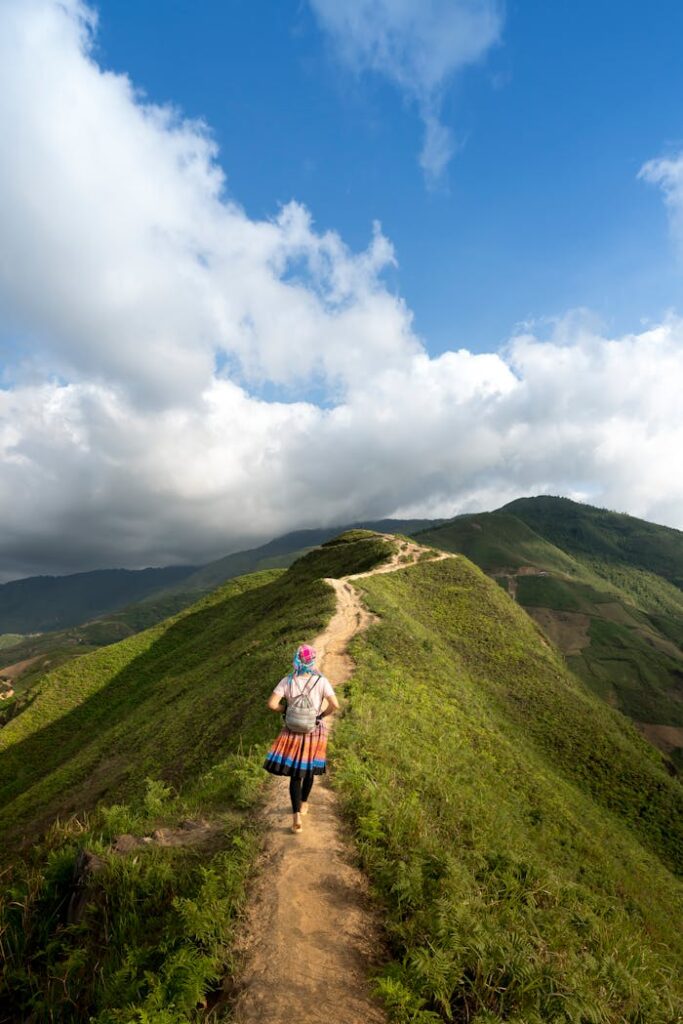 A lone hiker walking on a scenic mountain trail surrounded by lush greenery and clear blue skies.